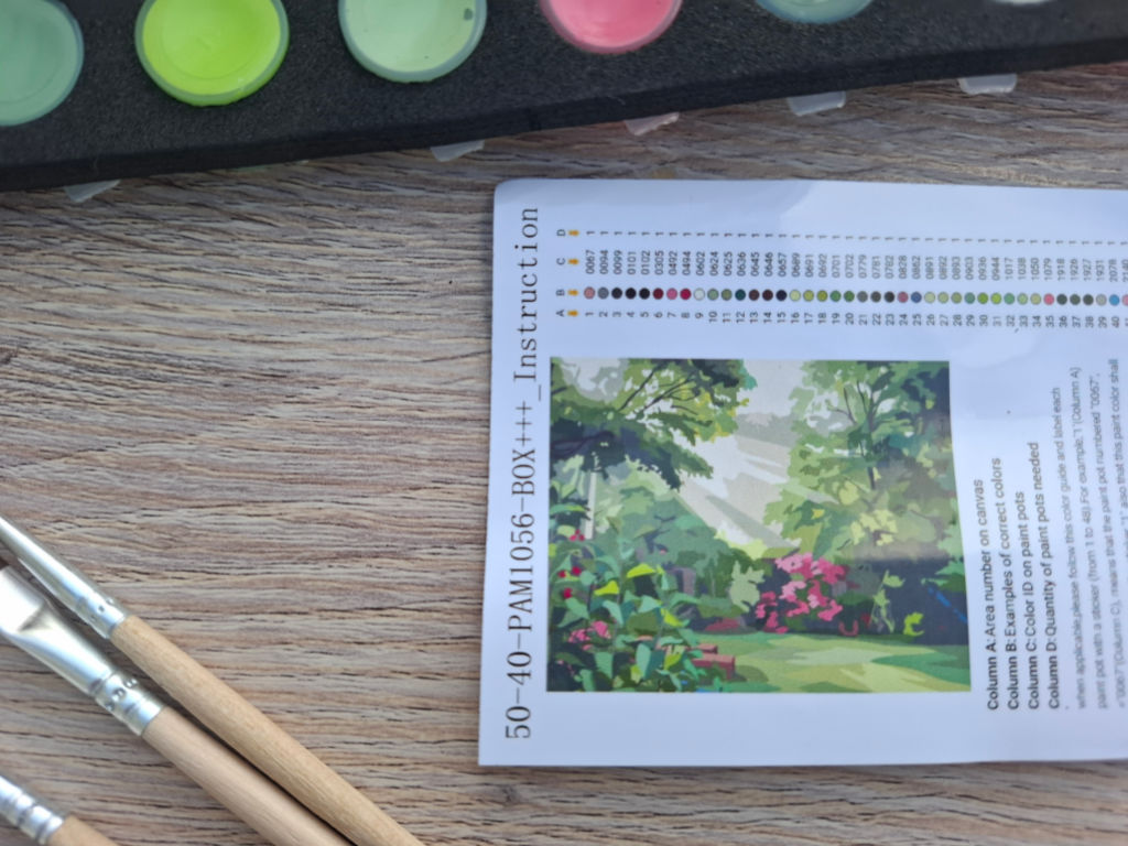 A paint-by-numbers instruction sheet shows a colourful garden scene beside brushes and paint pots on a wooden table, suggesting a calm, creative project.