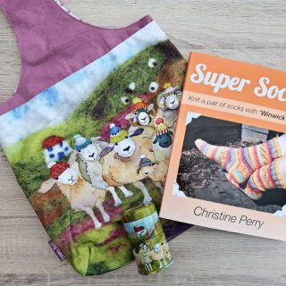 A colourful knitting project bag featuring playful sheep lies beside an orange “Super Socks” book on a wooden surface, creating a cheerful craft scene.